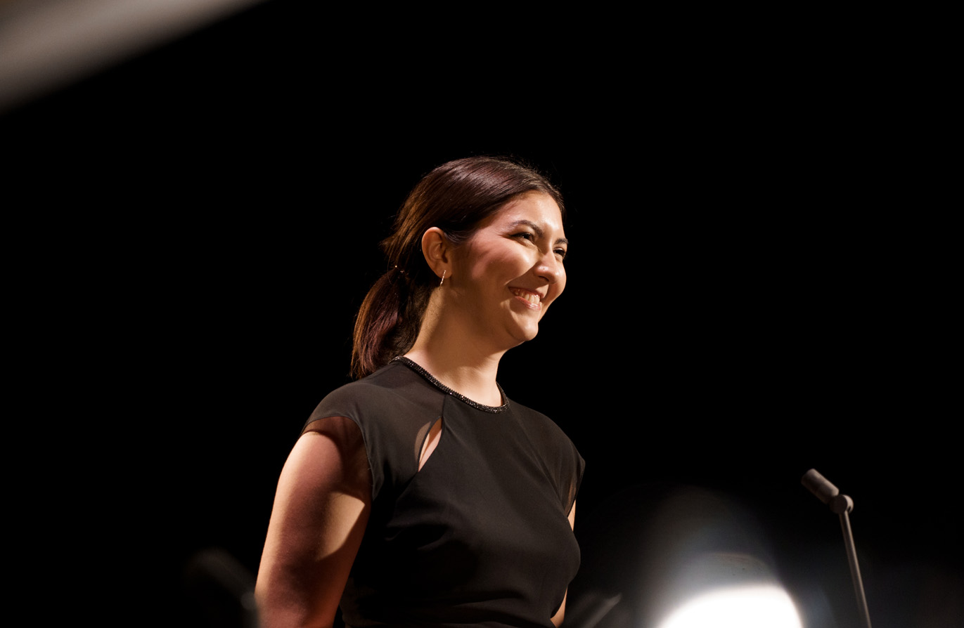 A female student, wearing smart attire, standing on stage and smiling at the audience.